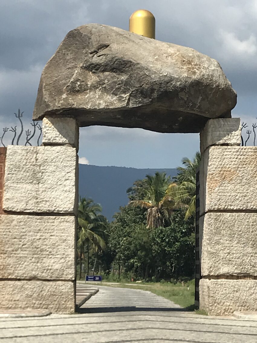 Stone archway with a large rock on top frames a scenic road and palm trees.